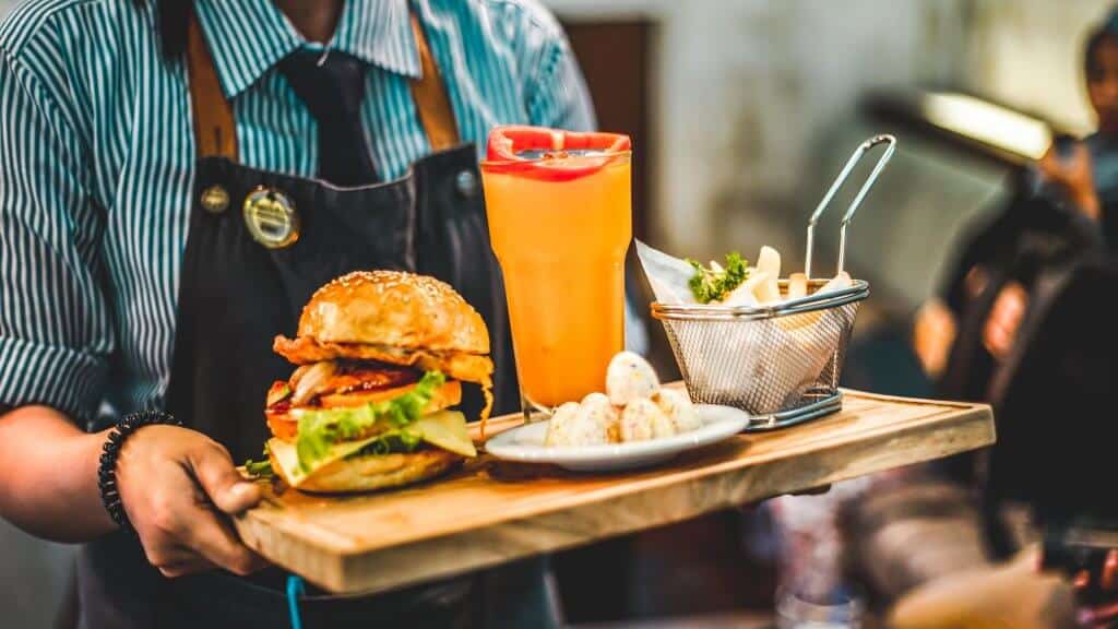 Waiter in blue striped shirt and apron carrying a wooden board with a burger, fries and drink