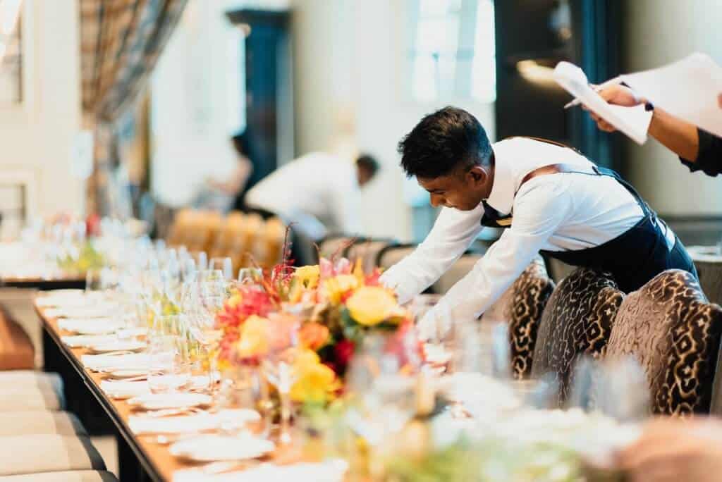 Waiter in a white shirt leaning over a long table with plates, flowers and glasses