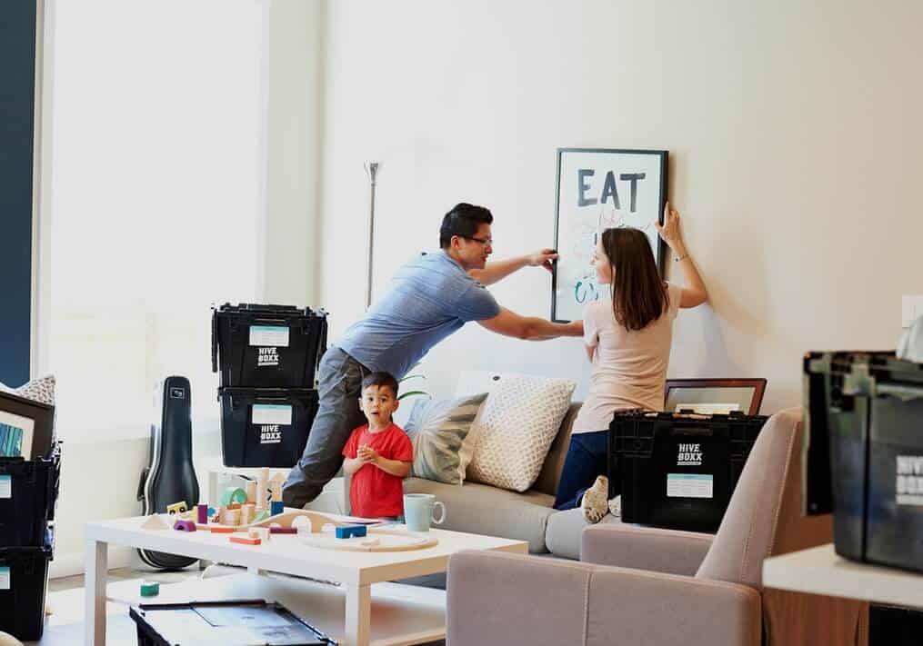 A man and a woman hanging a picture up in their new home as their child looks at the camera