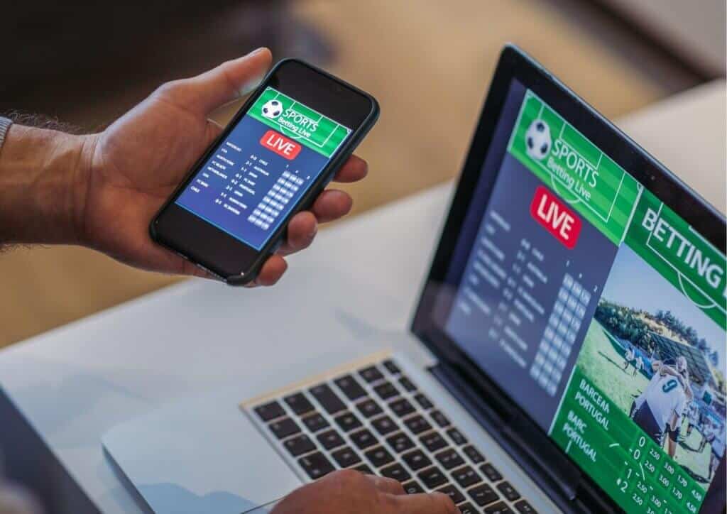 Hand holding a black phone next to a laptop - both screens show football related betting