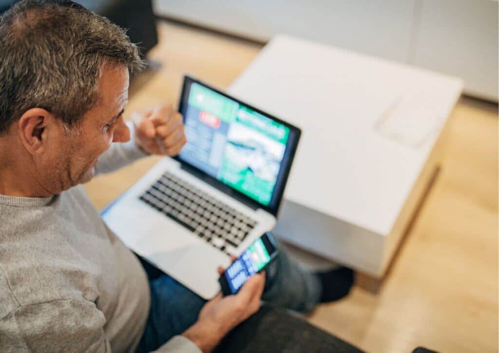 Man in a grey jumper holding a phone and sat with a laptop on his lap