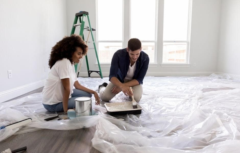 A young couple paint a blank room. There is a plastic floor covering, paint and a green ladder