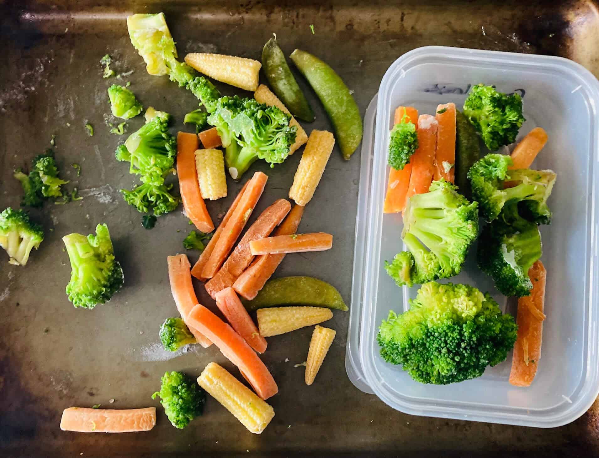 Frozen broccoli, carrots and babycorn, which are frozen on a bakinf tray. Some vegetables have been moved into a plastic container. 